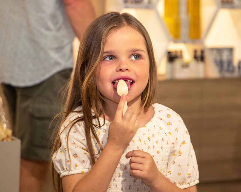 Girl in white dress with yellow daisy flowers, looking up licking spoon, honey products in background