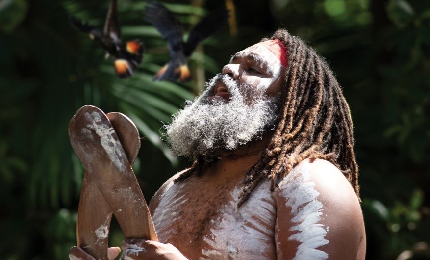 Man with long hair and beard bearing Australian Aboriginal markings on skin clapping boomerangs and singing while looking up