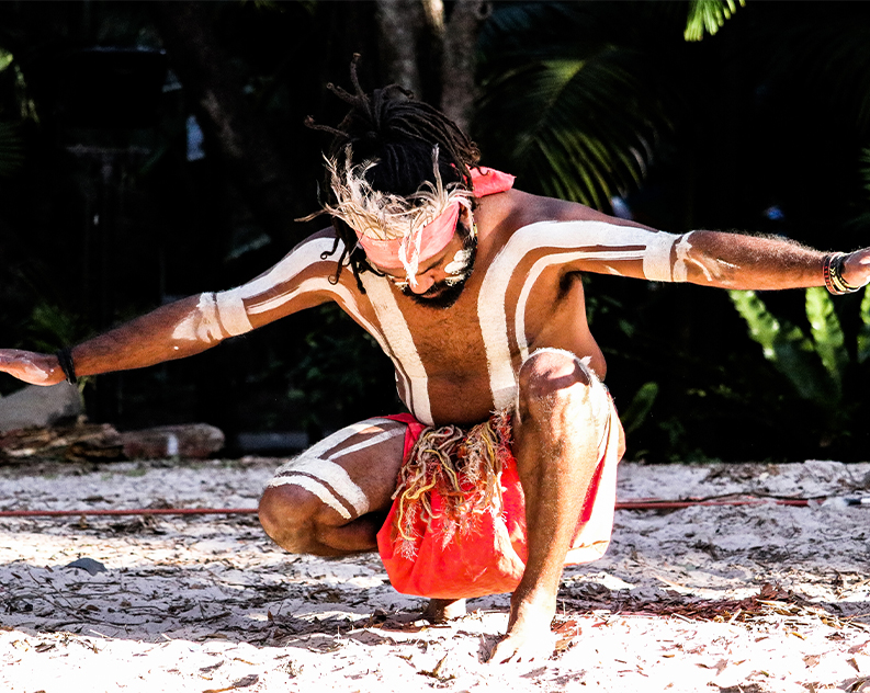 Man in traditional Australian Aboriginal clothing and markings crouching on sandy ground with arms outstretched in front of green trees