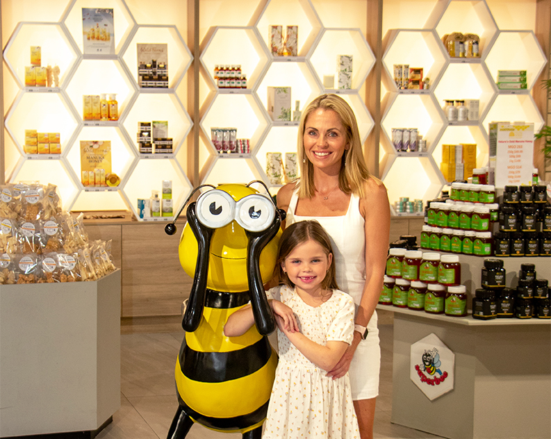 Mum and daughter smiling, posing next to giant bee statue inside honeyshop, stacks of honey products in honeycomb shaped shelves and on tables in background