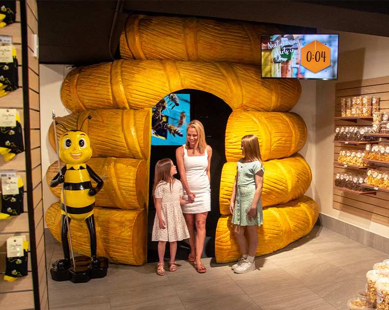 Mum and two daughters walking out of a honeycomb shaped area inside a honey shop, giant bee statue and stacks of honey products near them
