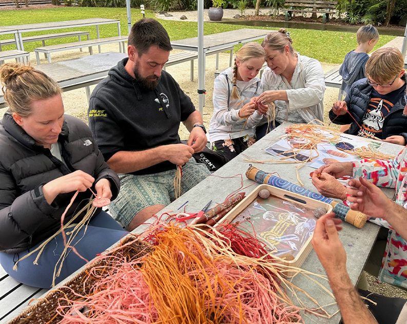 Group of people weaving, sitting around a table topped with colourful threads and a didgeridoo, surrounded by green grass