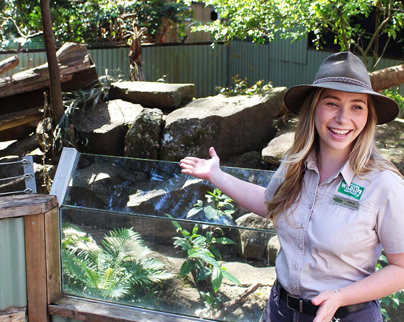 A woman in a khaki shirt and rancher hat smiling and standing in front of an animal habitat with lots of rocks and greenery, pointing at it
