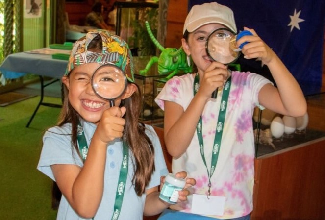 Two children, smiling and holding magnifying glasses, explore insects at a natural history museum.