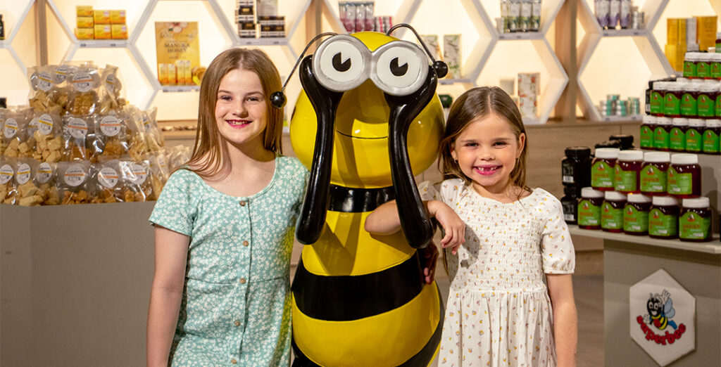 Two girls smiling, standing besides giant bee statue inside a shop, with stacks of packed honey products in background