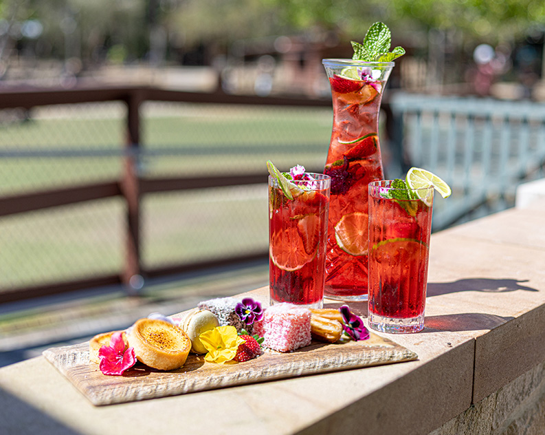 A mixed platter of colourful sweet treats placed on a concrete base with a jug and two glasses in the background filled with red liquid, fresh slices of lime and ice, topped with fresh mint, flowers and lime slices.