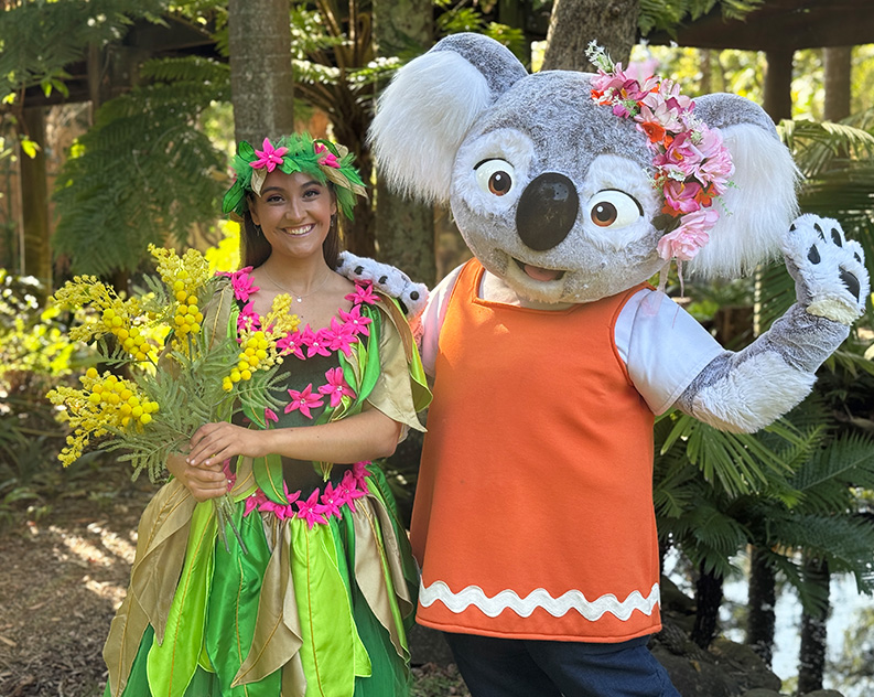 Smiling woman in a colourful fairy costume, holding yellow flower bouquet. She is standing next to a person in a life size koala costume, wearing an orange vest and pink flower crown.