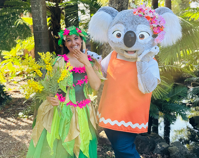 Woman in a colourful fairy costume giggling with her hand in front of her mouth, holding yellow flower bouquet. She is standing next to a person in a life size koala costume, wearing an orange vest and pink flower crown.