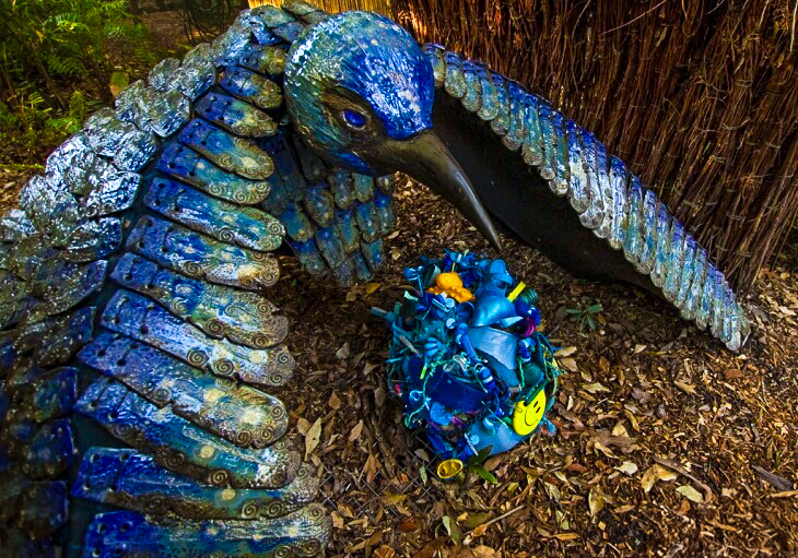 A large blue bird sculpture appears to protect a nest made of assorted blue objects and toys; SWELL Sculpture Festival logo is visible in the corner.