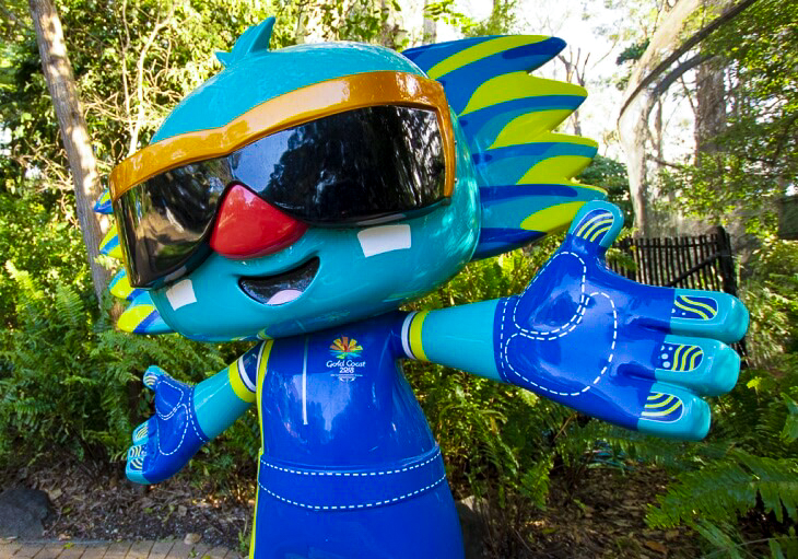 A colorful mascot statue with blue skin, large sunglasses, and spiky head feathers stands with arms outstretched in a garden setting.