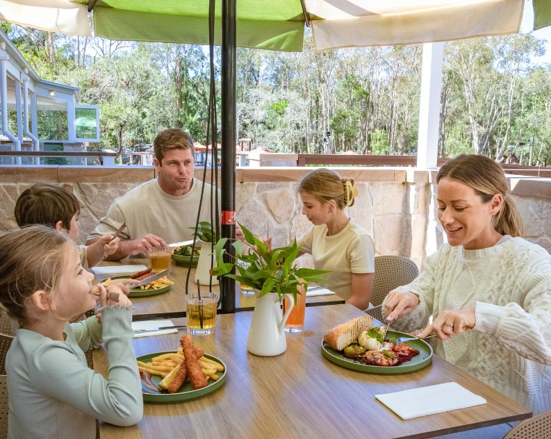 Family with three kids sitting at a table outdoors enjoying a beautiful lunch under a green and white umbrella, at a modern rustic venue