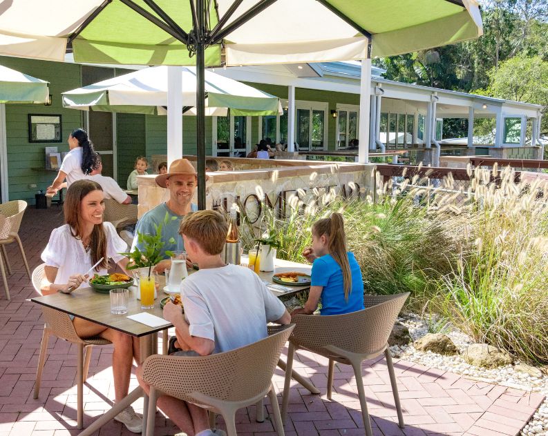 Family with two kids at an outdoor table, enjoying a nice lunch on a sunny day under a green and white umbrella