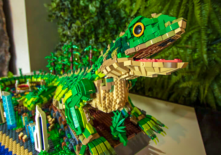 A large green lizard sculpture made from LEGO bricks is displayed on a table, surrounded by small LEGO trees and buildings.