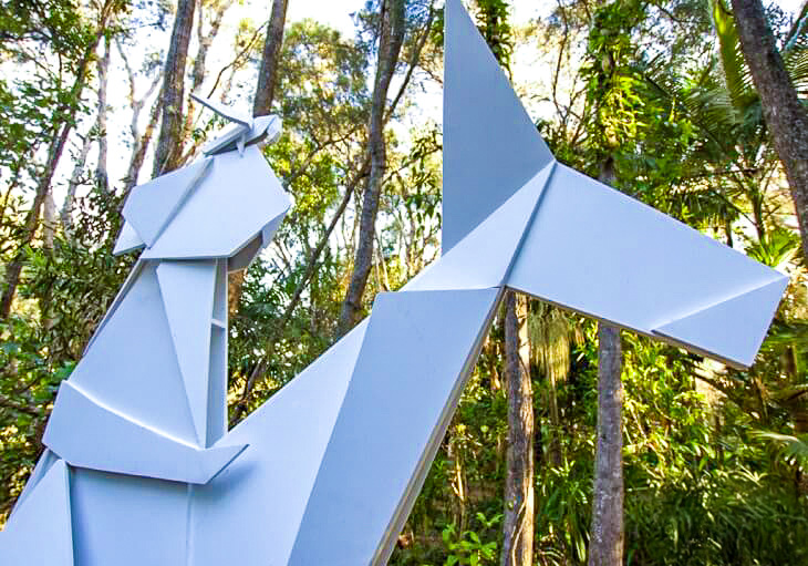 Large white geometric horse sculpture set among tall trees at an outdoor location, with SWELL Sculpture Festival logo in the corner.