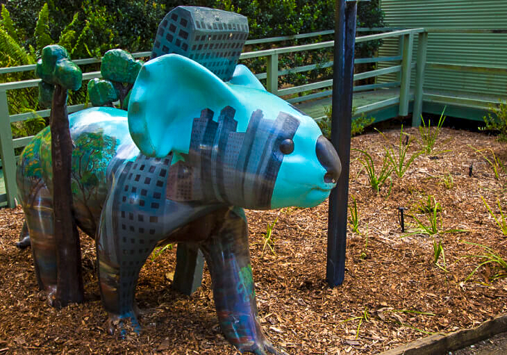 A painted koala statue featuring city buildings and trees on its body stands outdoors on wood chips near a railing.