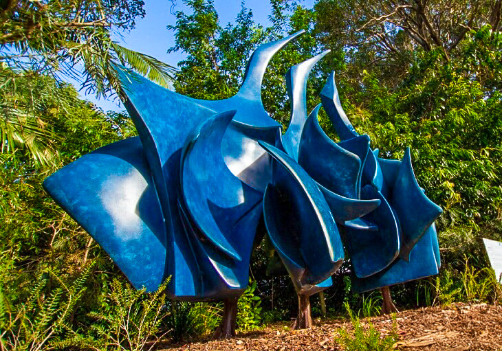 Abstract blue sculpture with curved, pointed shapes displayed outdoors among green trees and plants; SWELL Sculpture Festival logo at bottom right.