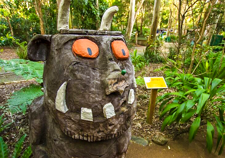 A large sculpture of a creature with orange eyes, white horns, tusks, and a wide mouth stands on a garden path surrounded by greenery and a yellow sign.