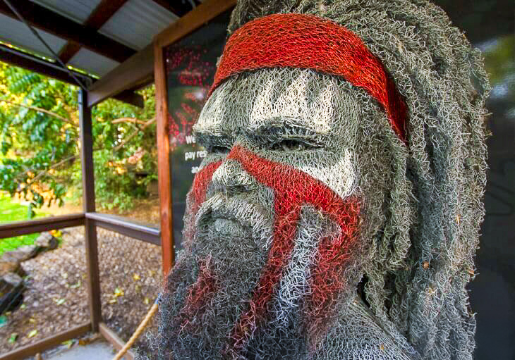 A wire mesh sculpture of a persons head and shoulders with red and white face paint, displayed at the SWELL Sculpture Festival.