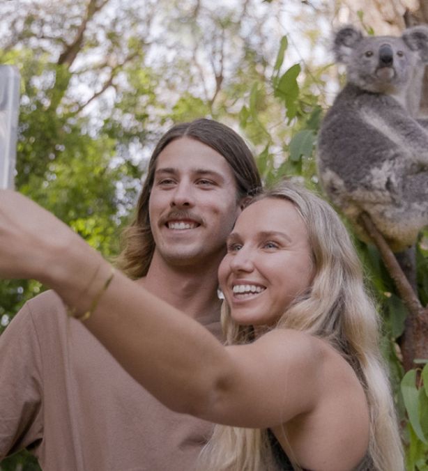 A young couple taking a selfie in front of a Koala in a tree behind them, very close to them.