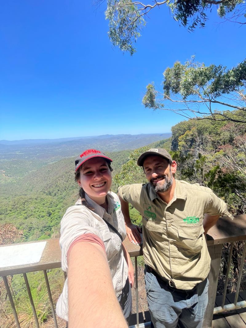 Man and woman in khaki clothing wearing caps taking a selfie on a viewing platform outlooking endless forests and a steel blue sky