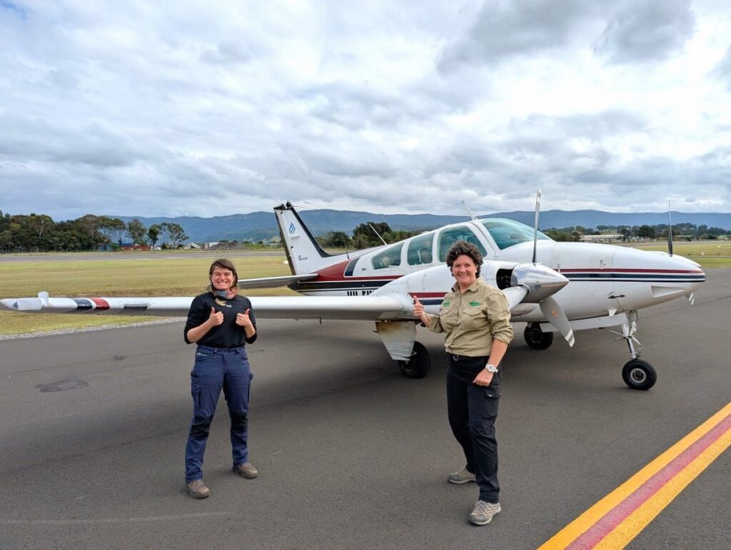 Two women in neutral outdoor clothing giving the thumbs up, standing in front of a small plane on a plane strip. Inside the plane, two more women look out the window smiling. In the background, there are mountain ranges and a large grass field, the sky is cloudy.