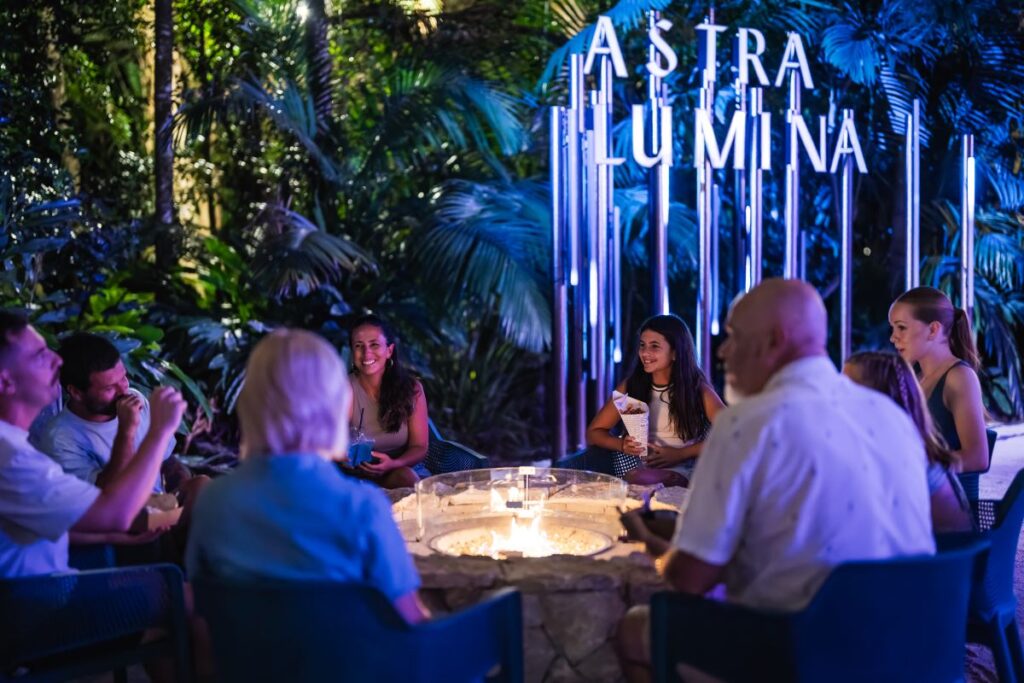 Group of people sitting around an artificial fire, laughing and talking to each other while some are holding a bag of hot chips. A large illuminated sign saying Astra Lumina is visible behind them