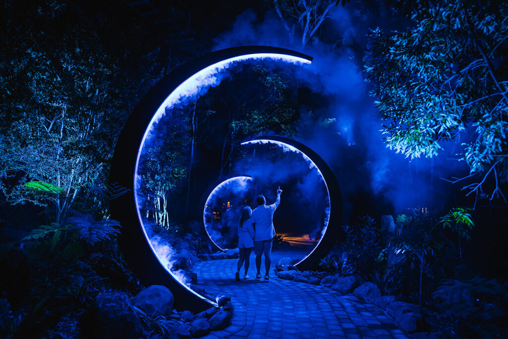 Couple walking through illuminated blue arches in a forest at night.