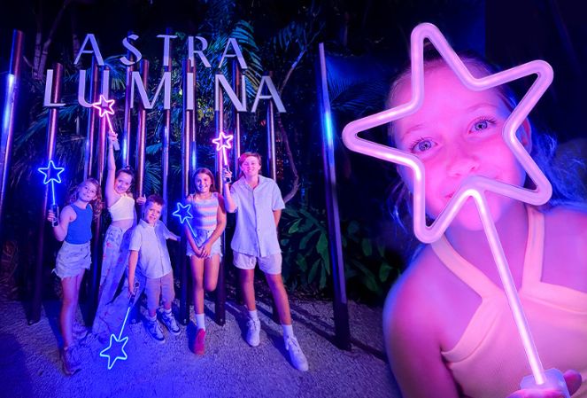 A girl holding up a glowing star lit up in purple colours. Next to her are five children with glowing stars as well, posing in front of a large illuminated photo prop with a sign saying Astra Lumina