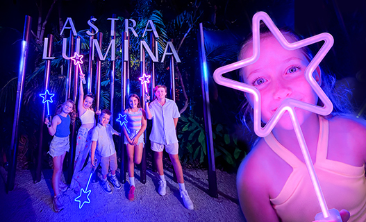 A girl holding up a glowing star lit up in purple colours. Next to her are five children with glowing stars as well, posing in front of a large illuminated photo prop with a sign saying Astra Lumina