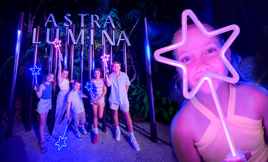 A girl holding up a glowing star lit up in purple colours. Next to her are five children with glowing stars as well, posing in front of a large illuminated photo prop with a sign saying Astra Lumina