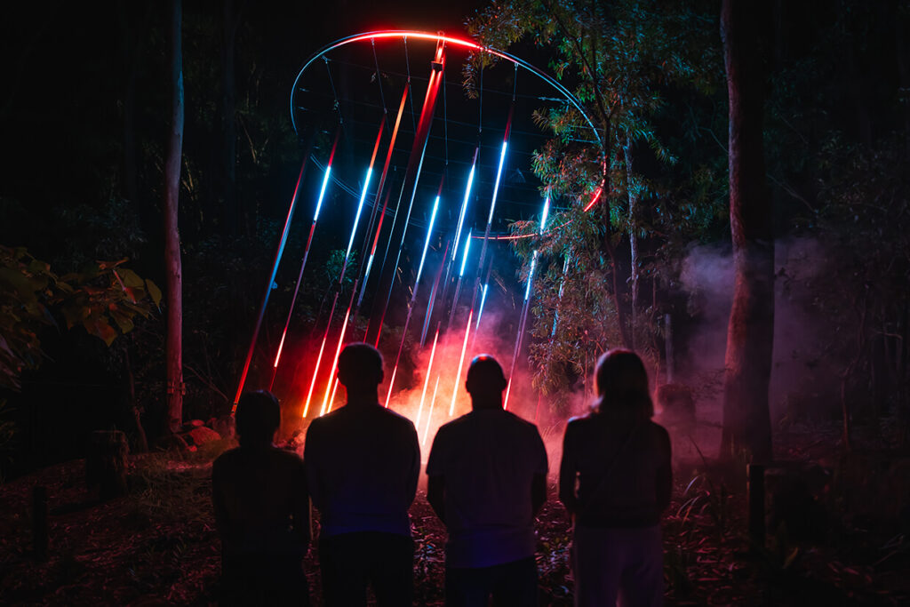 Silhouettes watching colorful light installation in woods.