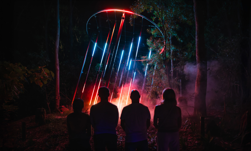 A light show playing at night with fog and different colours, set amongst natural bushland. Visible is the silhouette of a family of four watching the show.