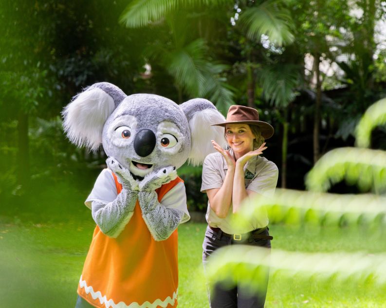 A person in a life size Koala costume wearing navy pants and an orange vest standing next to a female wildlife keeper wearing khaki outdoor clothes and a cowboy hat. Both are posing and smiling, standing amongst beautiful green trees and grass