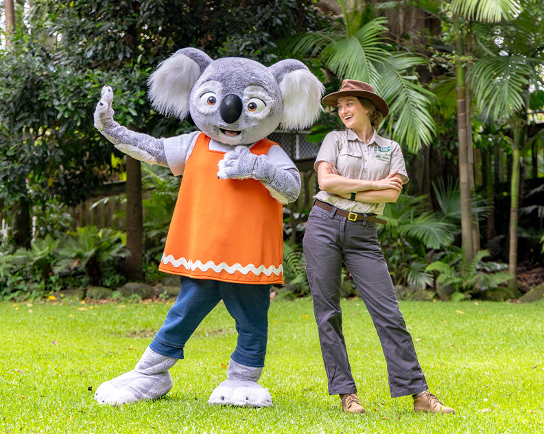 A person in a life size Koala costume wearing navy pants and an orange vest standing next to a female wildlife keeper wearing khaki outdoor clothes and a cowboy hat. Both are posing and standing on lush green grass with green large trees in the background