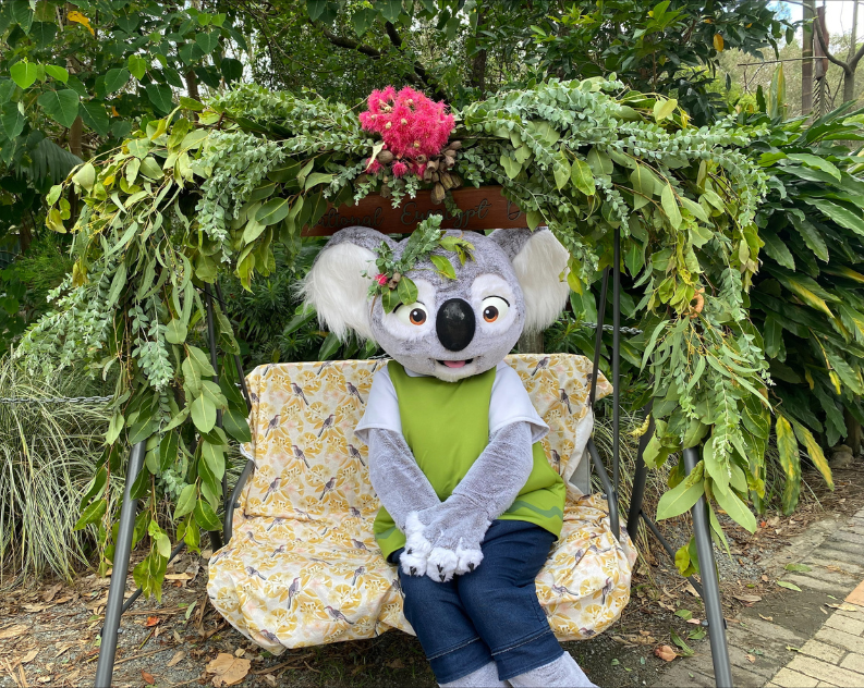 A person in a life size koala costume sitting on a bench under a lush green native flower arch