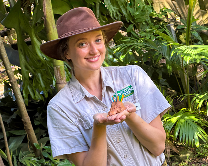A woman wearing a brown Stenson hat and a khaki t-shirt holding up a small echdina made out of clay and craft materials. She is standing in front of tropical green trees 