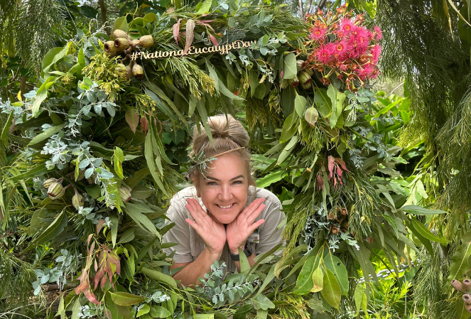 Smiling blonde woman poking her head through the middle of a beautiful lush green eucalypt wreath with native flowers