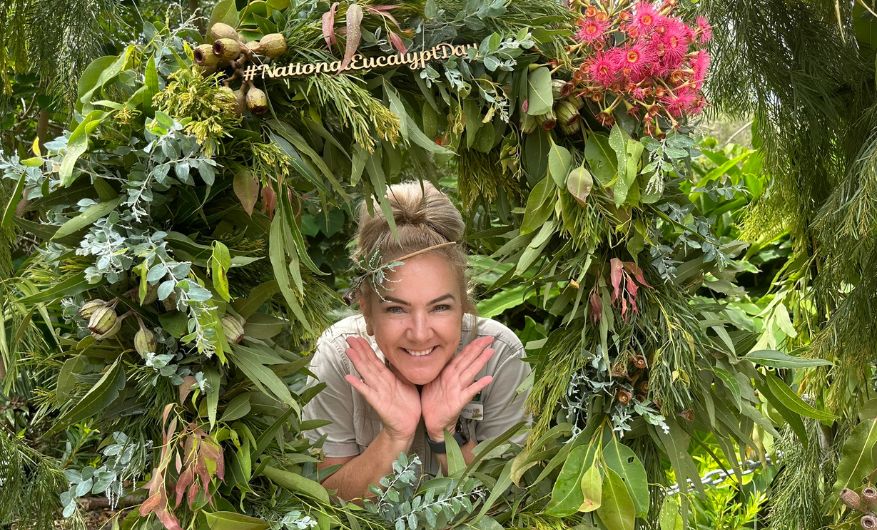 Smiling blonde woman poking her head through the middle of a beautiful lush green eucalypt wreath with native flowers