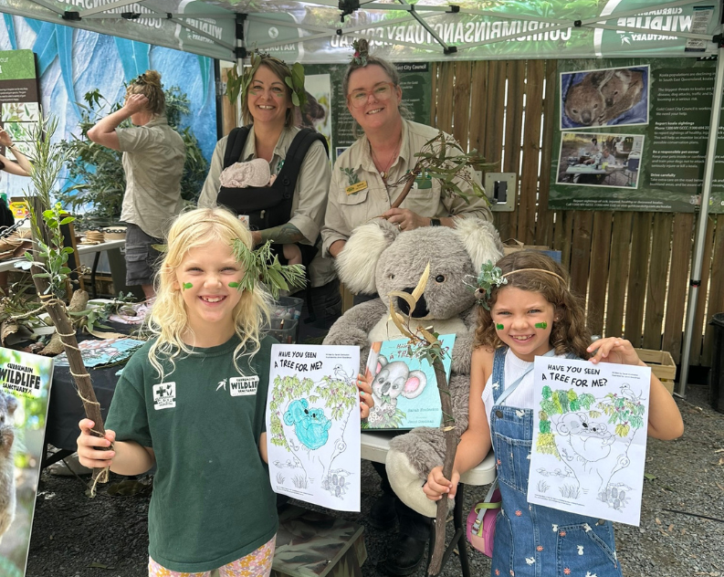 Two young girls smiling with eucalypt leaves in their hair, holding up a eucalypt and koala drawing as well as a wooden stick decorated with gum leaves. Behind them are two female wildlife keepers at a craft stand