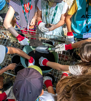 A group of kids around a bucket filled with soil, wearing garden gloves and holding a small shovel