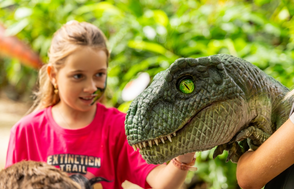 A real looking dinosaur hand puppet close-up with a girl in the background patting it. She has a green dinosaur painted on her face and is wearing a pink t shirt with dinosaurs on it. Around them are green trees