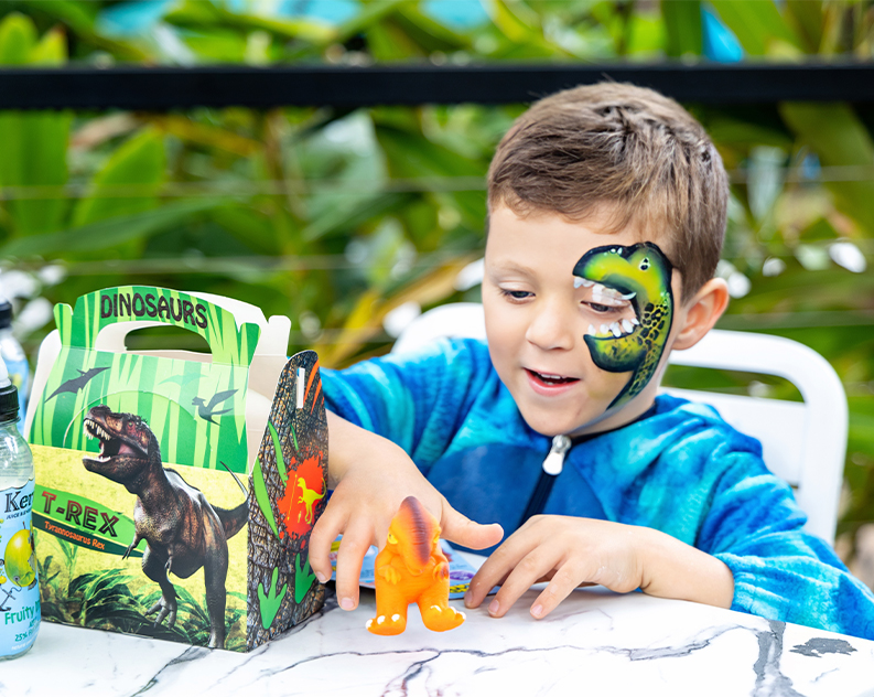 A young boy in a blue dinosaur suit playing with an orange dinosaur toy while sitting at a table that has a dinosaur meal pack on it. His face has a green dinosaur painted on it