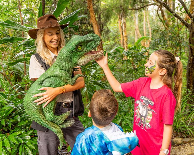 A green dinosaur hand puppet held by a blonde woman in khaki clothing and a stetson hat with a young boy and girl in dinosaur outfits patting and looking at the puppet