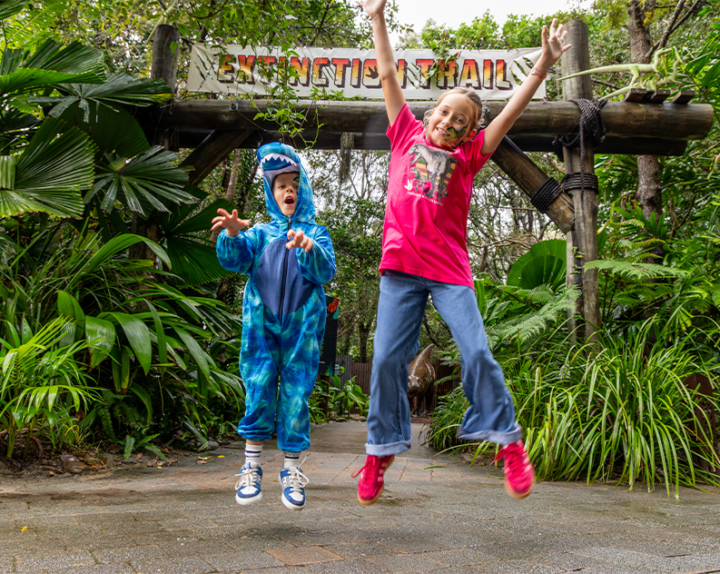 Girl wearing pink dinosaur t shirt and sneakers and blue jeans, and boy wearing a blue dinosaur overall and white sneakers, in mid air jumping up in front of a wooden arch with a sign saying extinction trail