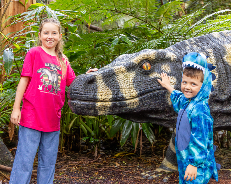 A young girl and boy wearing dinosaur outfits standing next to a giant lizard replica. Both are patting the lizard's snout and looking at the camera