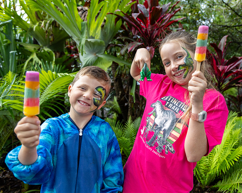 Girl and boy in dinosaur outfits holding a colourful ice cream and a small dinosaur toy. Both have a dinosaur painted on their face