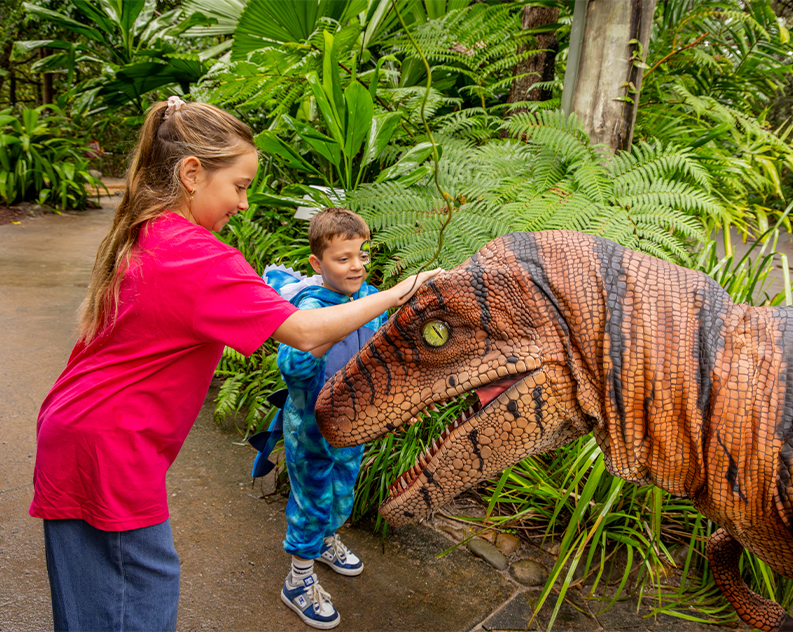 Girl wearing pink shirt and blue jeans with boy in blue dinosaur overall suit, both patting a real looking dinosaur. They are on a concrete pathway, around them are large tropical green trees