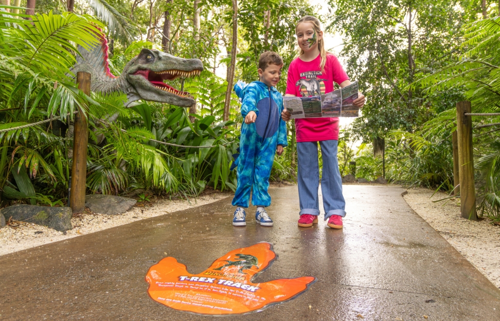 A boy in a blue dinosaur suit and girl in pink dinosaur shirt standing in front of a large orange dinosaur footprint on the pathway. The girl is looking at a map and the boy is pointing to the footprint. In the background is a large dinosaur replica peaking out of green bushland