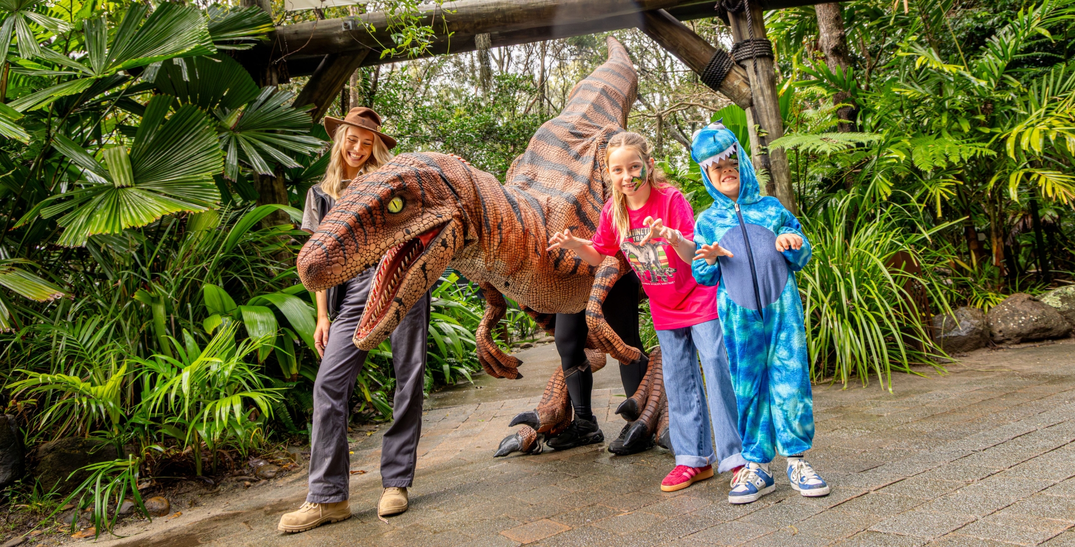 A girl in a pink dinosaur shirt and jeans and a boy in a blue dinosaur suit making raw gestures right next to a life-size dinosaur replica. A young woman wearing khaki outdoor clothing and a stetson hat is smiling standing on the other side of the dinosaur. They are in an outdoor area with tropical trees in the background