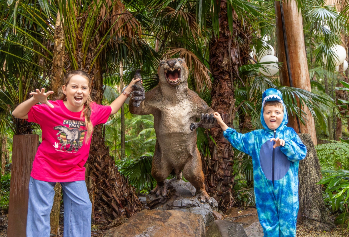 A girl in a pink dinosaur shirt and jeans and a boy in a blue dinosaur suit making raw gestures right next to a life-size dinosaur replica. They are in an outdoor area with tropical trees and greenery in the background